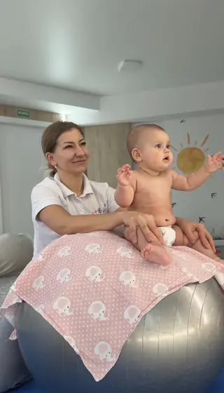 Femeie în tricou alb sprijină un bebeluș cu scutec, așezat pe o minge mare de gimnastică acoperită cu o pătură roz cu elefanți albi.