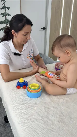 Adultă în tricou alb, așezată lângă un bebeluș pe o suprafață albă; îi întinde un inel colorat, iar lângă ei sunt jucării de sortat.