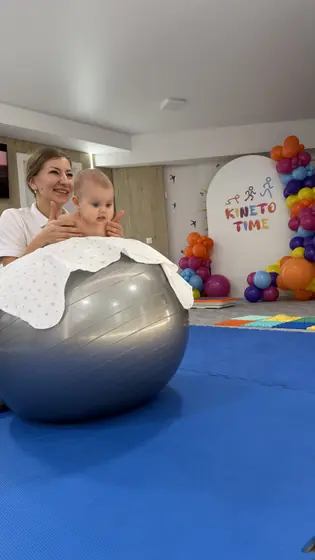 Femeie în tricou alb ținând un bebeluș așezat pe o minge mare gri acoperită cu o pânză albă, în sala cu covor albastru și baloane colorate.
