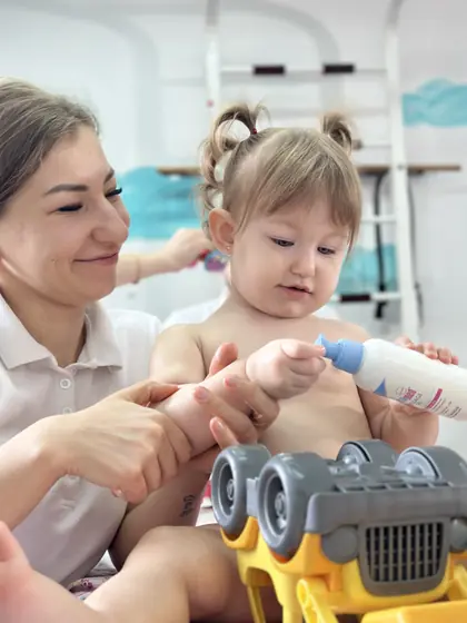 Femeie în tricou alb, zâmbind, ține de braț o fetiță cu două cocuri care se uită la un biberon cu capac albastru; în prim-plan, un camion de jucărie.