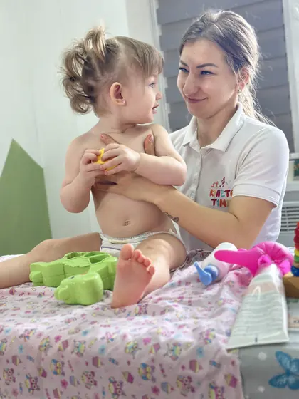 Femeie adultă ținând în brațe un copil mic așezat pe masa de masaj, cu jucării colorate în prim-plan, într-o cameră luminoasă.
