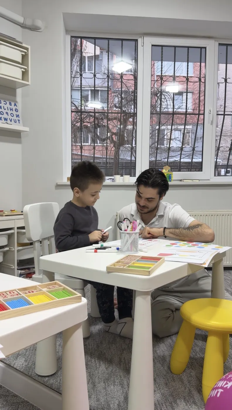 Copil și adult la o măsuță albă, lucrând cu foi colorate și cuburi de lemn