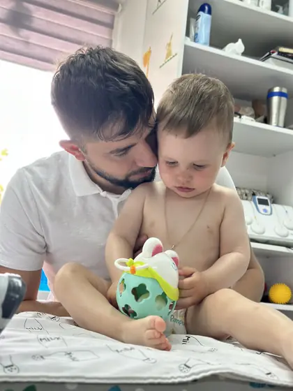 Un barbat in tricou alb tine in brate un copil mic asezat pe o patura, cu o jucarie turcoaz in maini, in fata unui raft cu aparat de fizioterapie.