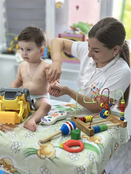 Fetiță în scutec pe masa cu jucării colorate; o femeie în tricou alb îi susține brațul, lângă fereastră.