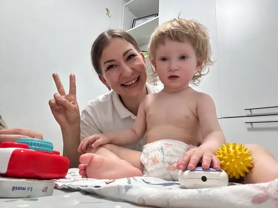 Femeie adulta asezata pe saltea, facand semnul V cu mana, langa un copil mic in scutec, inconjurati de jucarii colorate in cabinetul de kinetoterapie.
