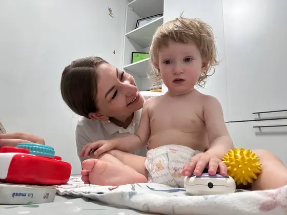 Copil mic așezat pe o pătură pe saltea, lângă o femeie adultă zâmbitoare; în față, o cutie roșie de jucării, o minge galbenă cu țepi și un obiect alb.