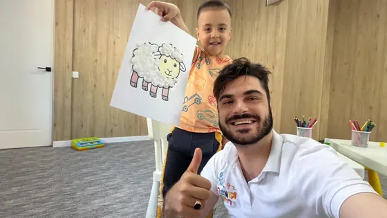 Barbat cu barba in tricou alb face selfie cu degetul mare ridicat, langa un baiat care tine sus un desen cu o oaie colorata, intr-o sala cu pereti de lemn.